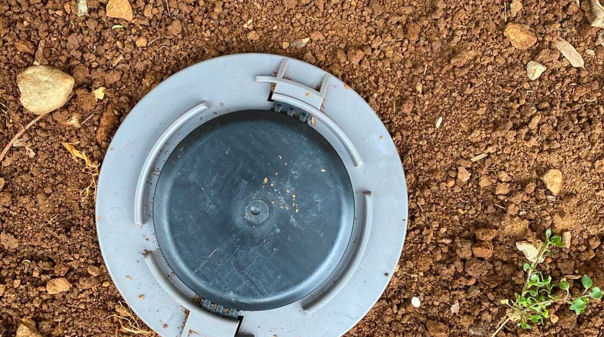 An in-ground termite bait station set flush with the soil beside a house, its cap closed.